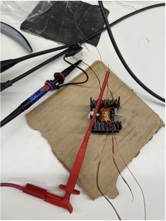 Hand-wound E-core flyback transformer on a lab workbench with oscilloscope probes and test leads attached